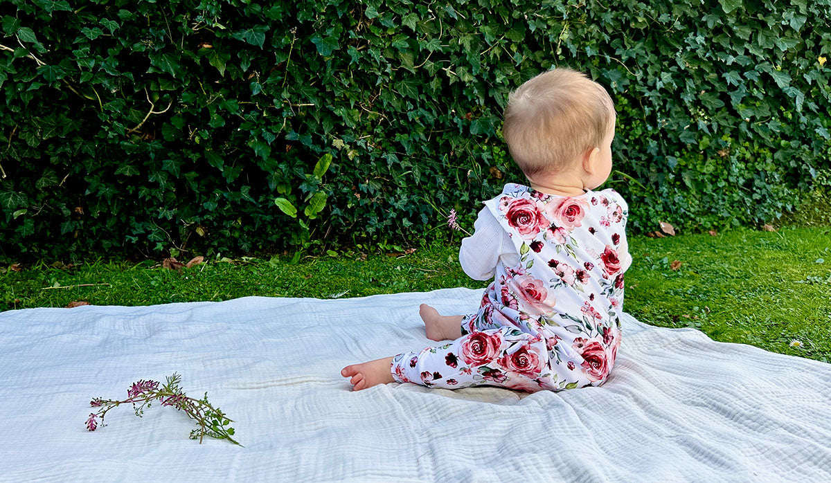Baby wearing Willa rose romper and white long sleeved top sitting on picnic blanket outside with a flower in hand